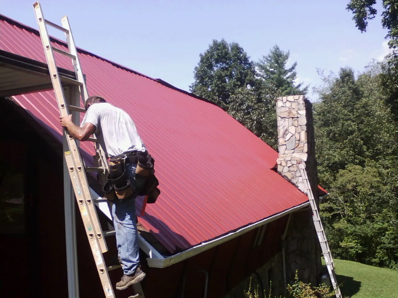 Roofer on a ladder beside a red metal roof and stone chimney — Soffit & Fascia Repair by Roofing Brothers in Kinsman